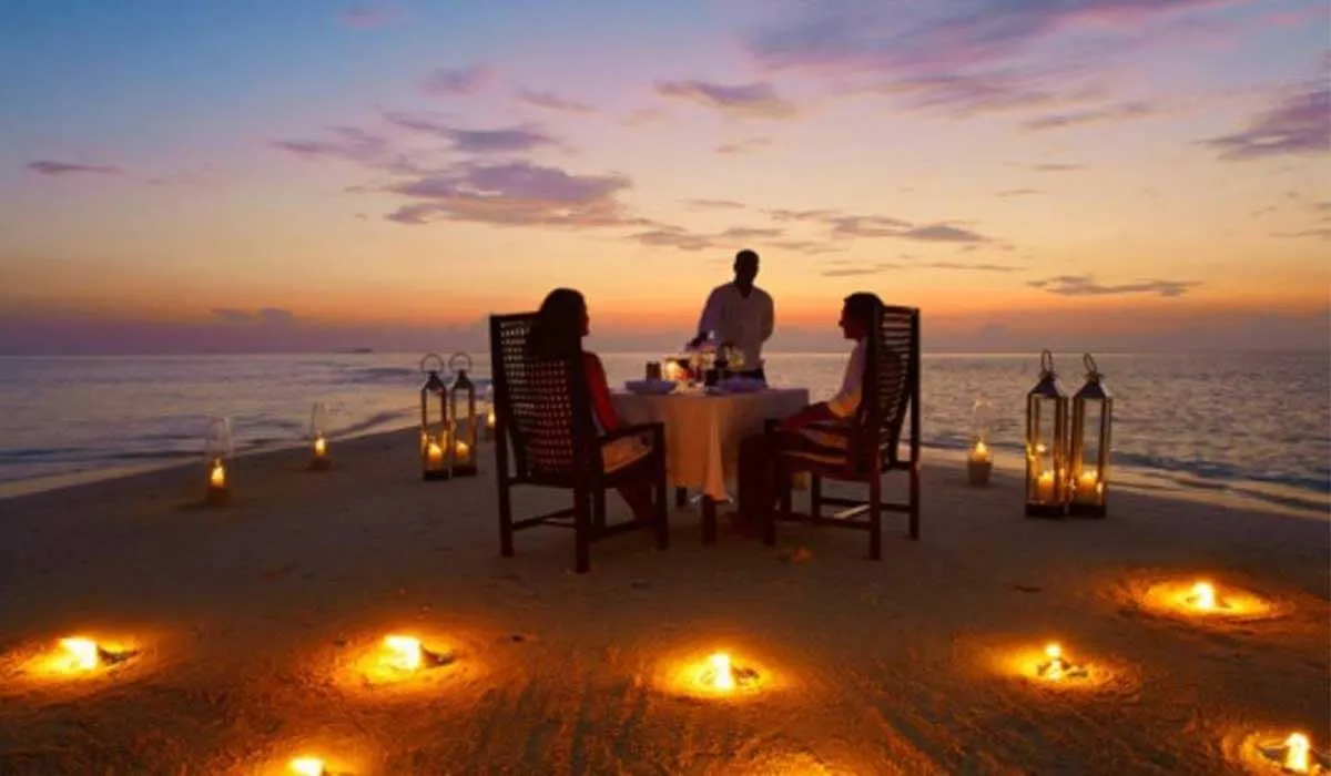 Couple dining on the beach at sunset, circled by candles, with a waiter serving