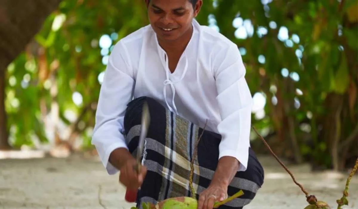 Smiling man in a white shirt and sarong opening a coconut on the beach