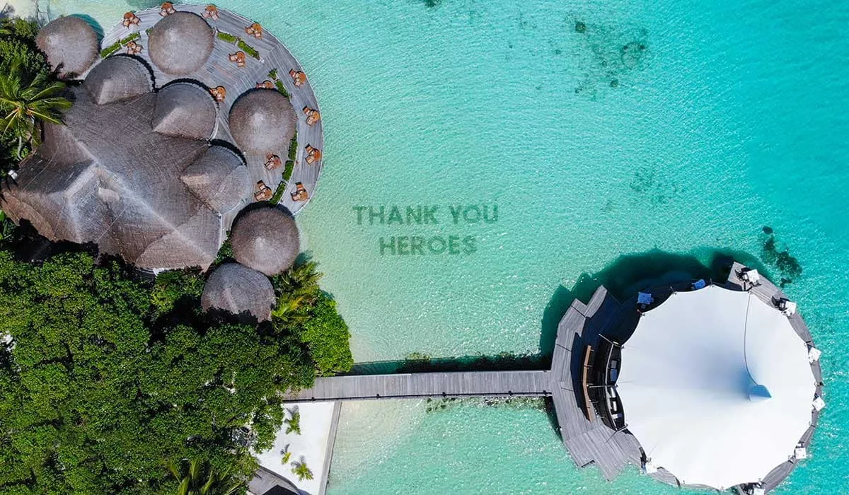 Aerial view of thatched villas and a white tent, with "Thank you heroes" spelled out in the lagoon