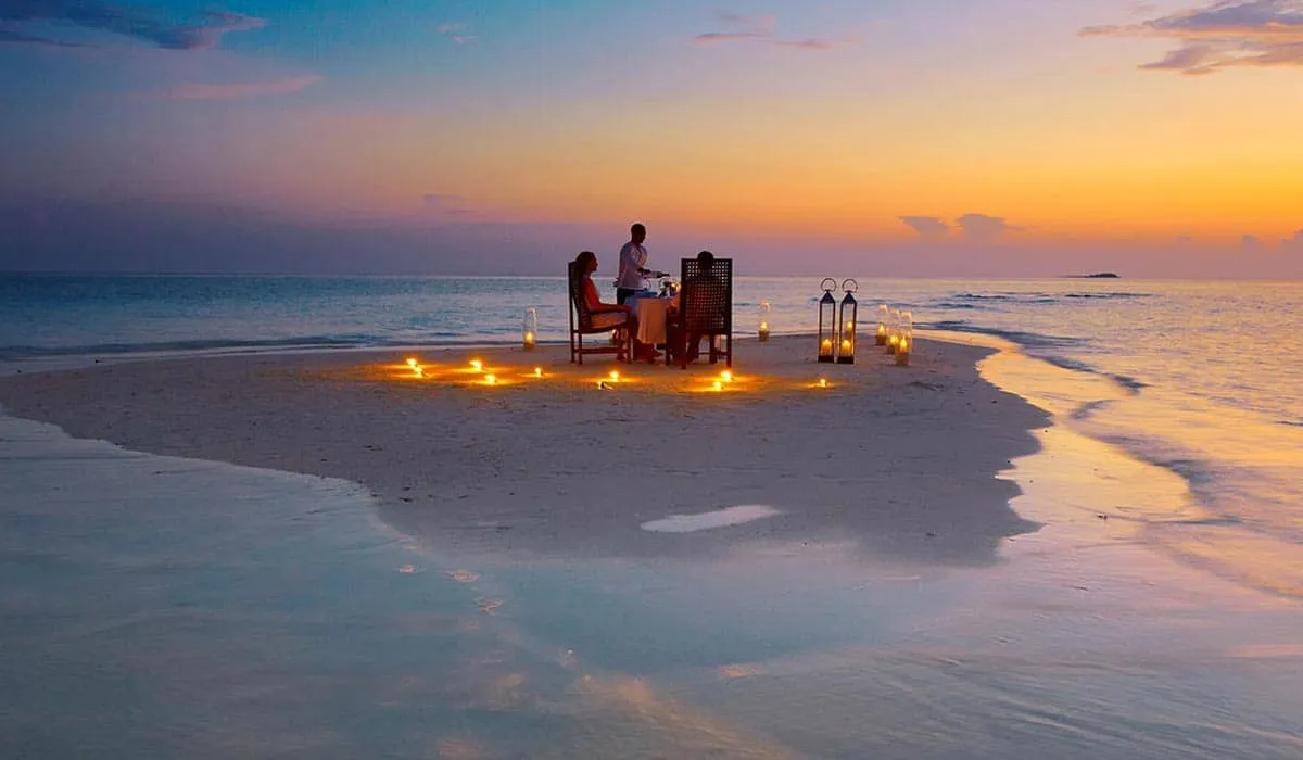 Couple dining on a candlelit sandbank at sunset, with a waiter serving