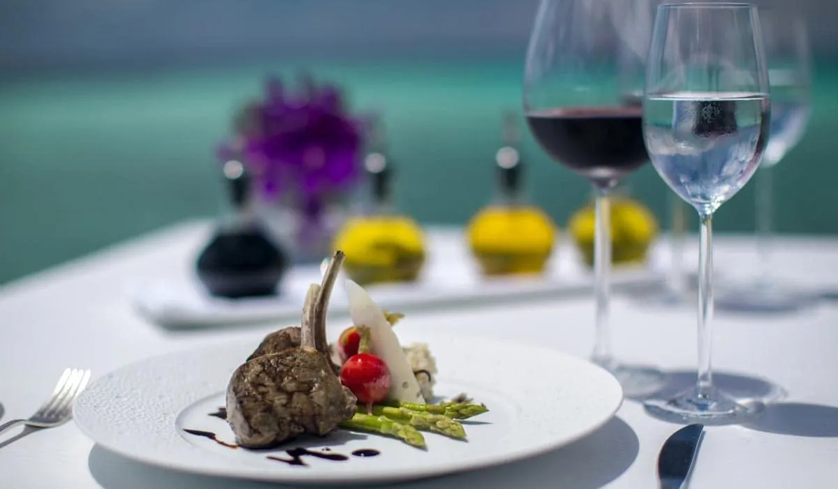 Plated meat dish with vegetables beside wine glasses on a white table