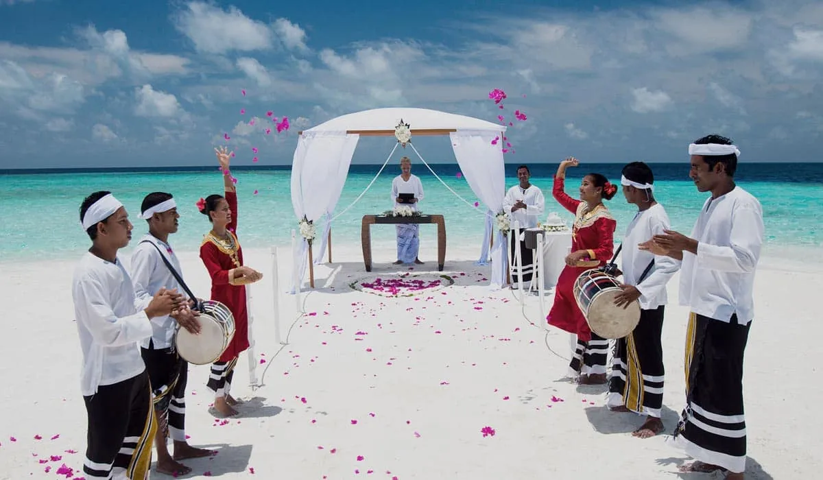 Couple at a beach wedding under a white canopy, with traditional drummers and dancers