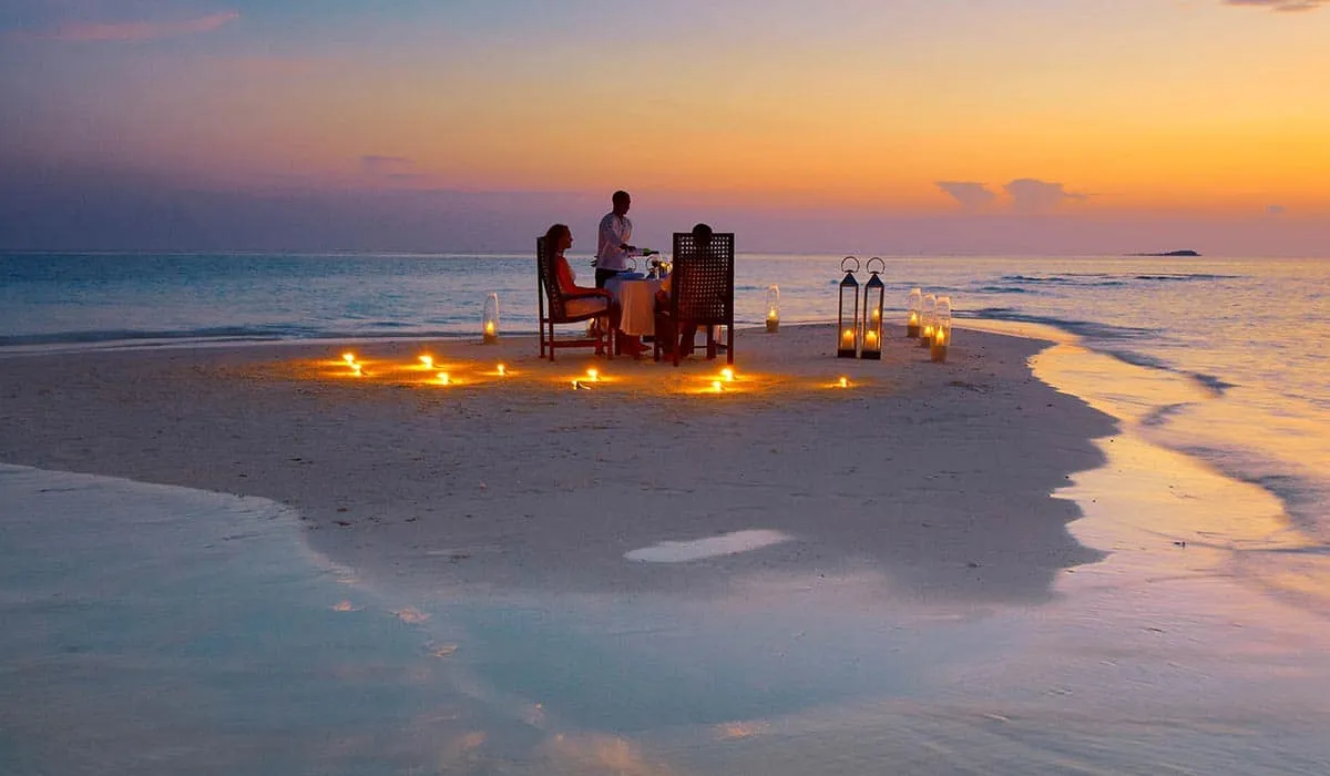 Couple dining on a candlelit sandbank at sunset, with a waiter serving