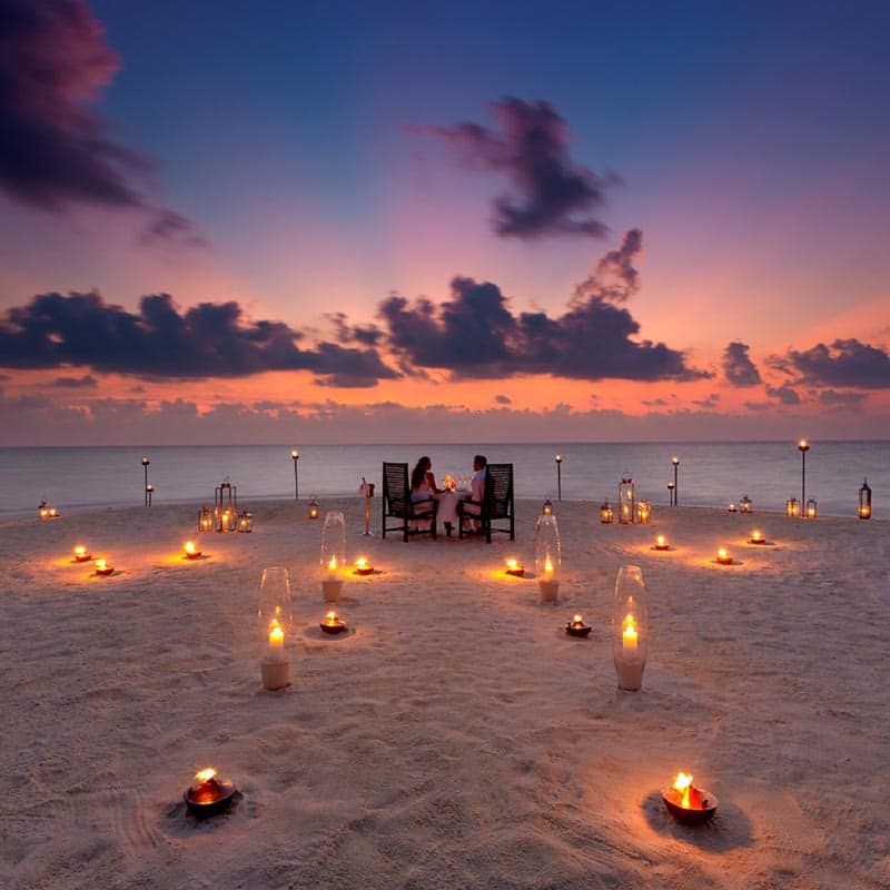 Couple dining on the beach at sunset, surrounded by candles in the sand