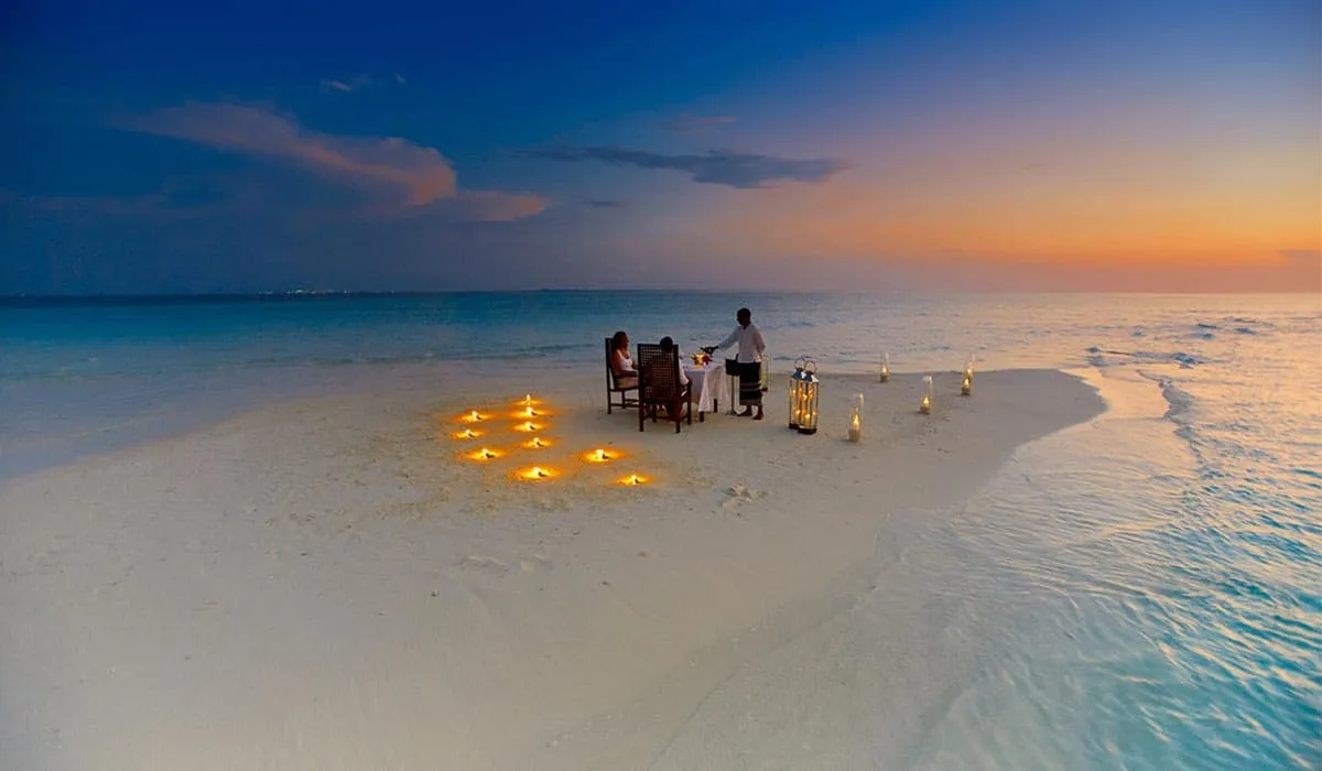 Couple dining on a candlelit sandbank at sunset, with a waiter