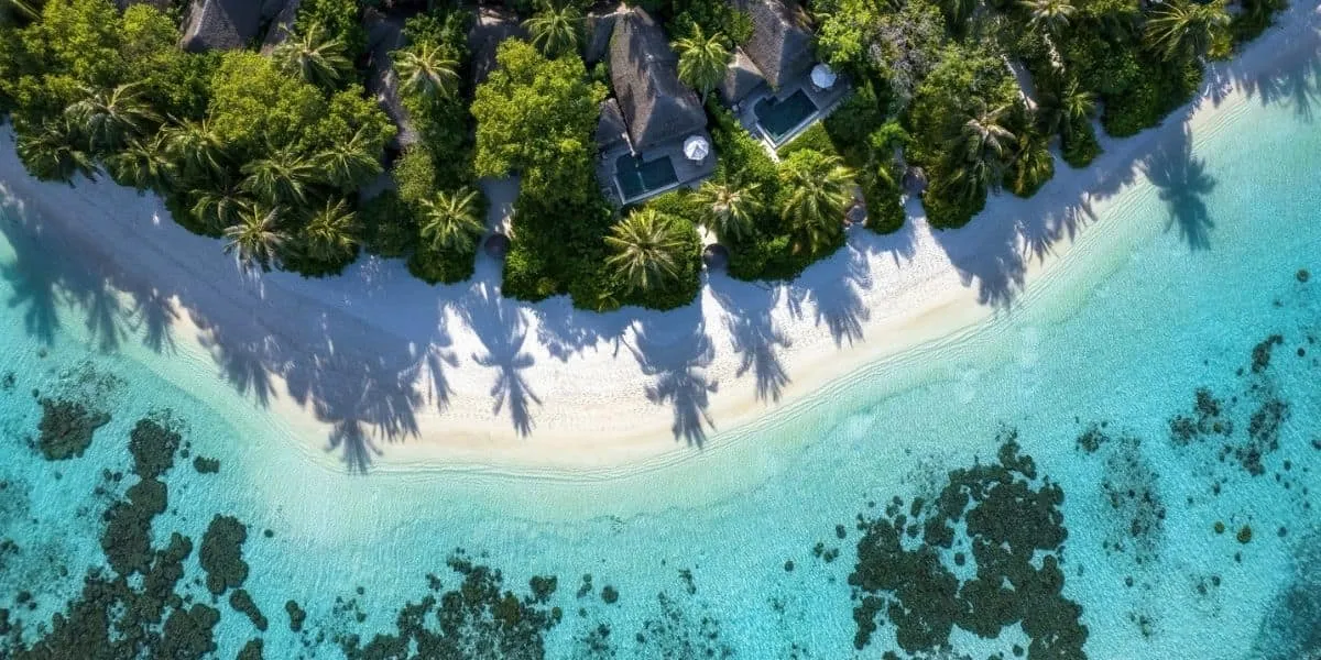Aerial view of a palm-fringed beach, villa roofs and clear lagoon