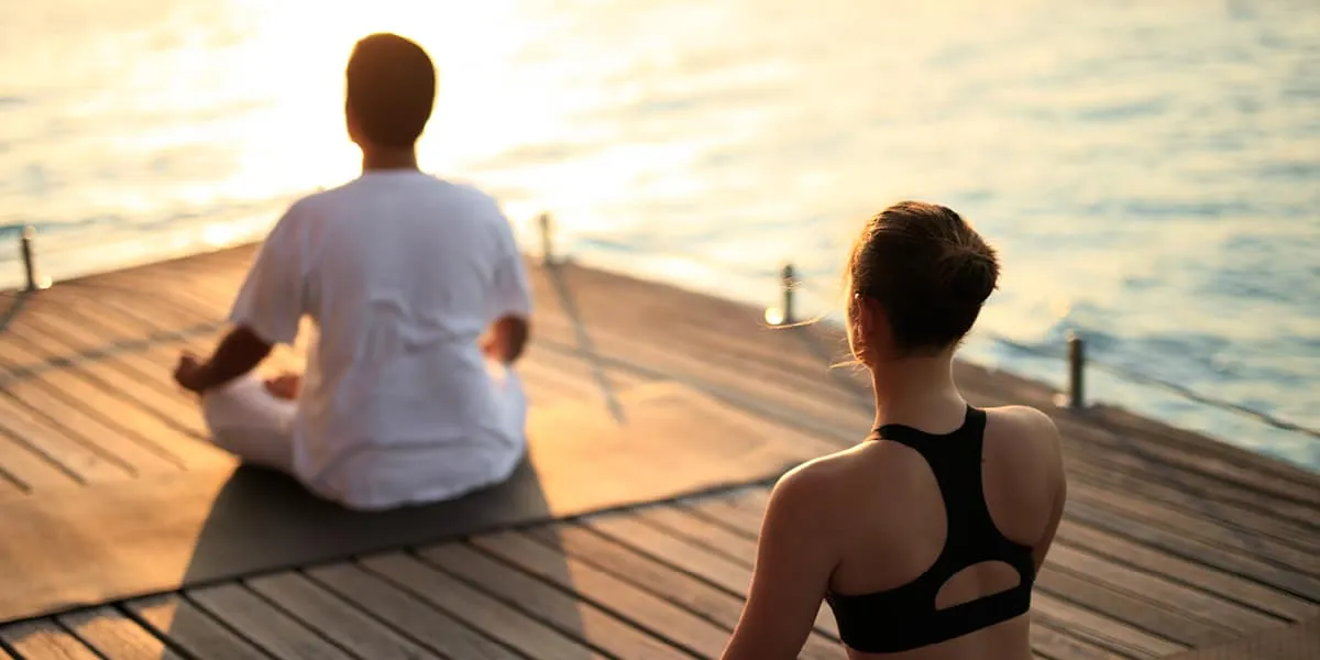 Two people meditating on a wooden deck overlooking the sea at sunset