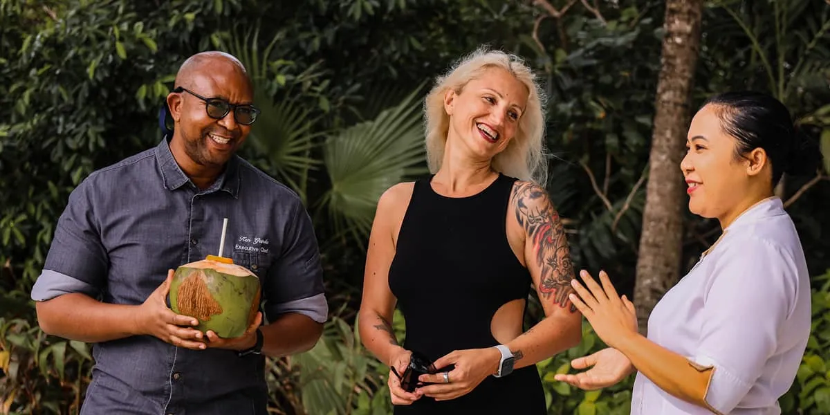 Three people laughing together in a garden, one holding a coconut