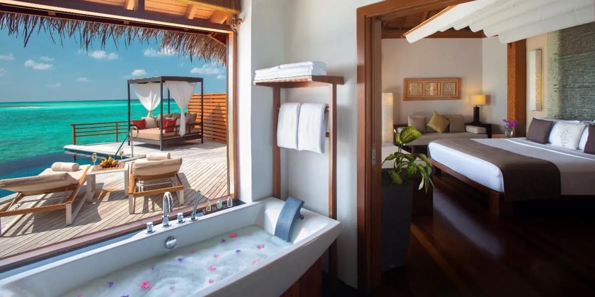 Water villa deck with a daybed and petal-filled bath, and a villa bedroom