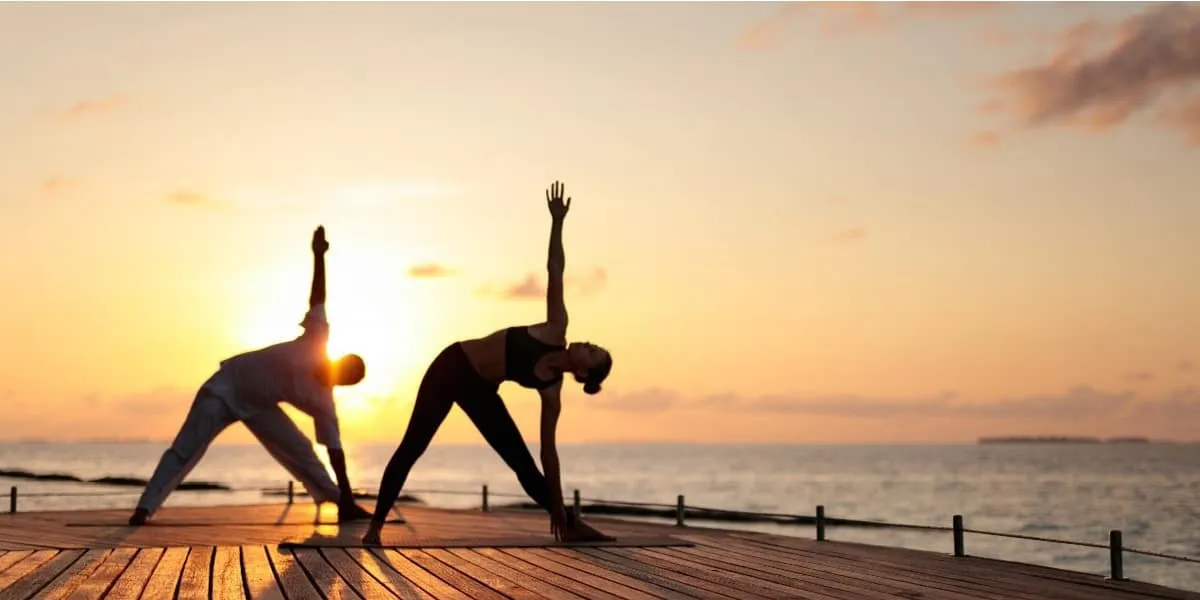 Two people in a triangle yoga pose on a deck at sunset