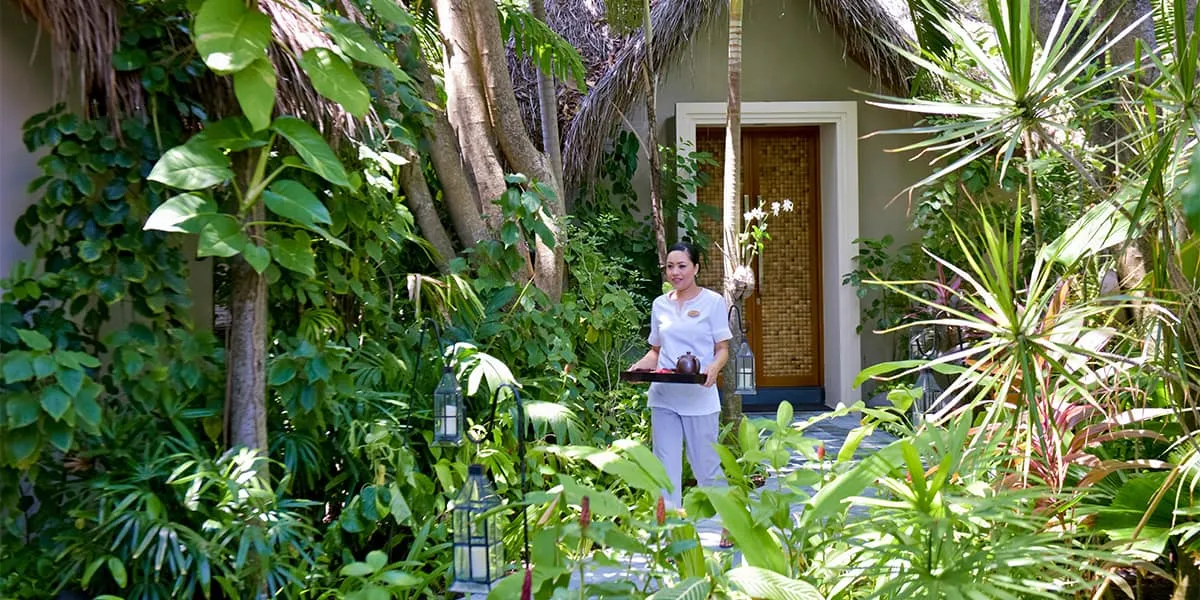 Therapist carrying a tray along a garden path outside a spa building