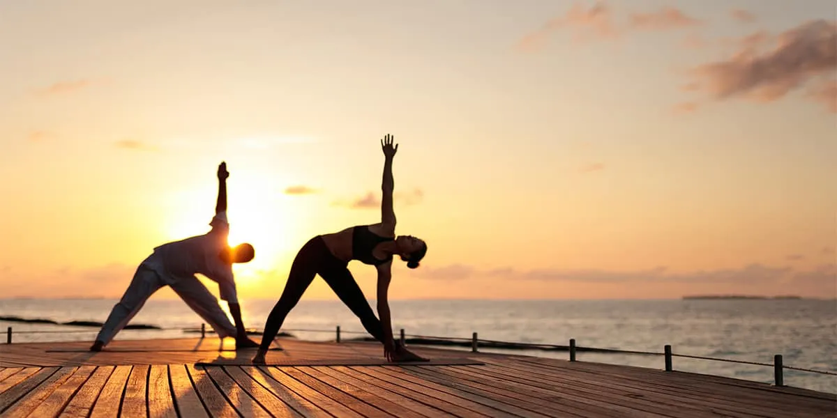 Two people in a triangle yoga pose on a deck at sunset