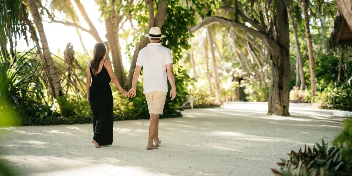 Couple walking hand in hand along a sandy path shaded by trees at Baros