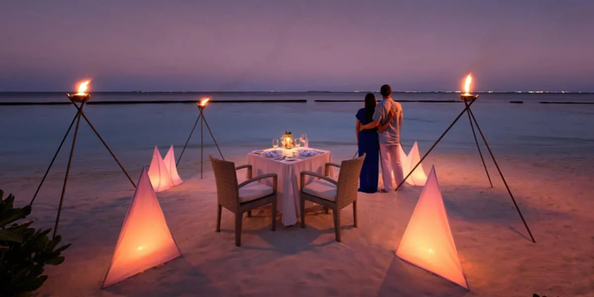 Couple beside a candlelit dinner table on the beach at dusk, lit by torches
