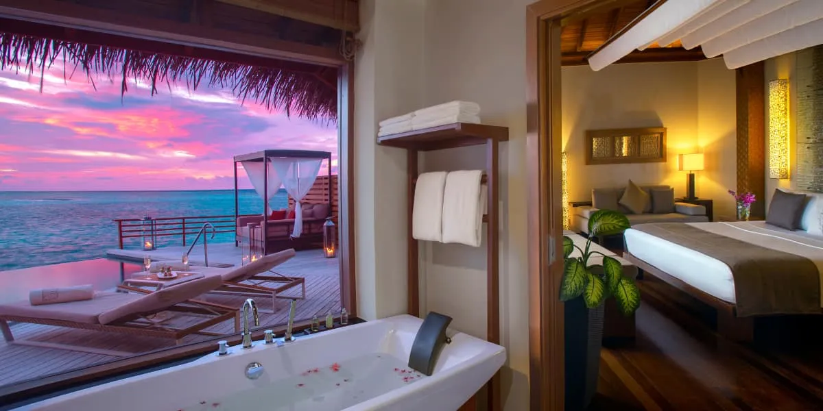 Water villa with an outdoor bathtub, bedroom and deck facing a pink sunset