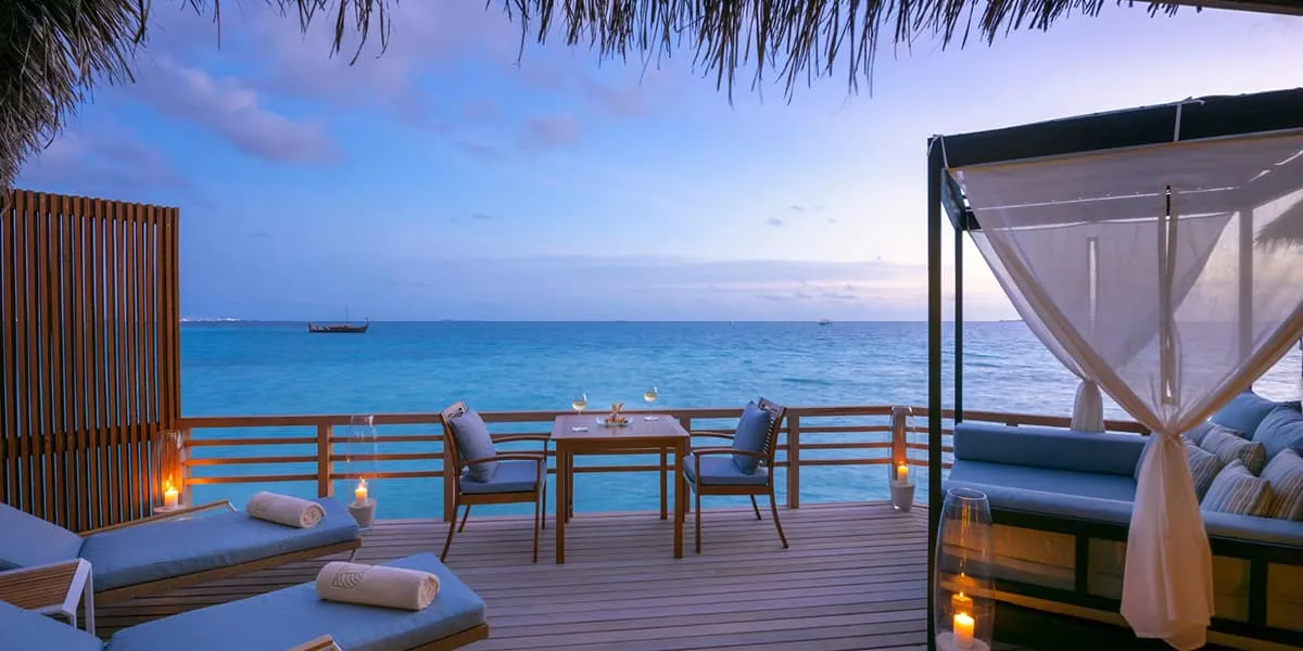 Water villa deck with a dining table, daybed and sea view at dusk