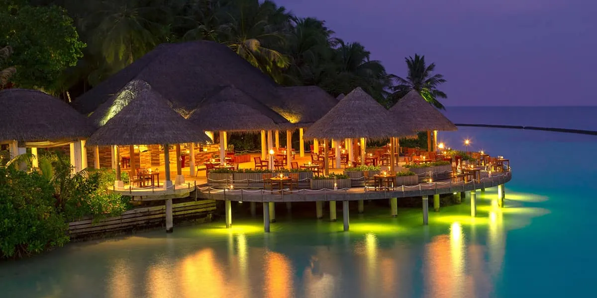 Thatched overwater restaurant lit up at dusk above the lagoon