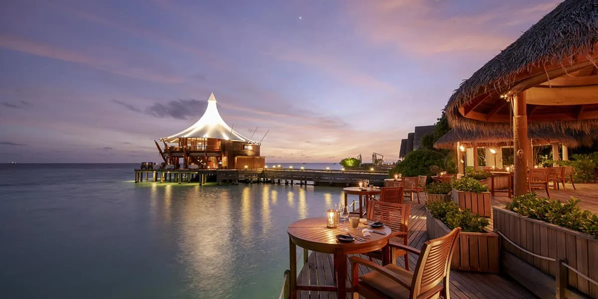 Candlelit restaurant deck over the water at dusk, with a lit pavilion beyond