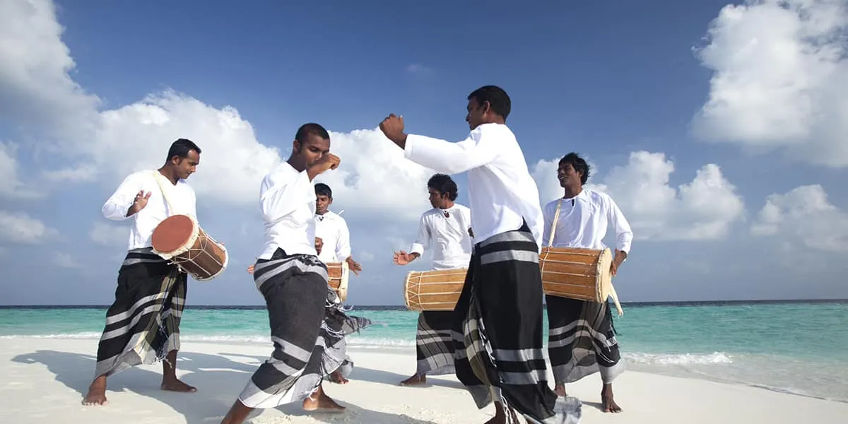 Men in white shirts and sarongs playing Maldivian drums on the beach