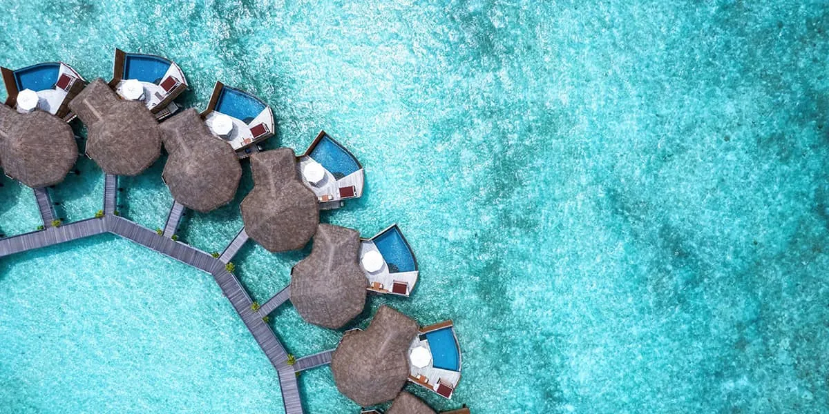 Aerial view of thatched water villas along a curved jetty over the lagoon