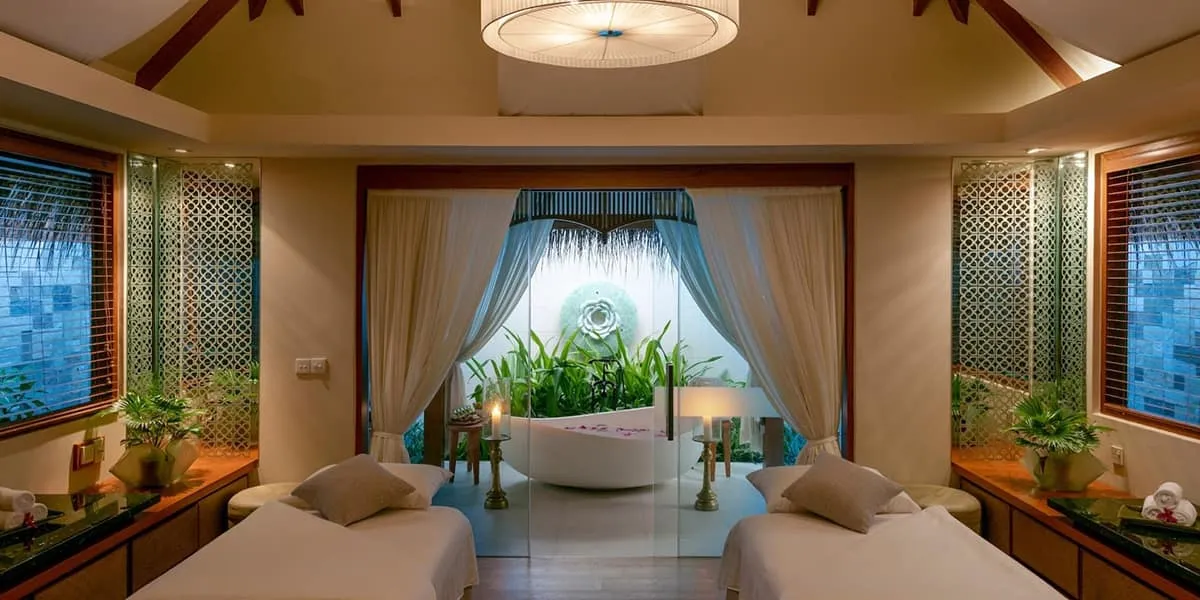 Spa treatment room with twin massage beds, a bathtub and tropical plants
