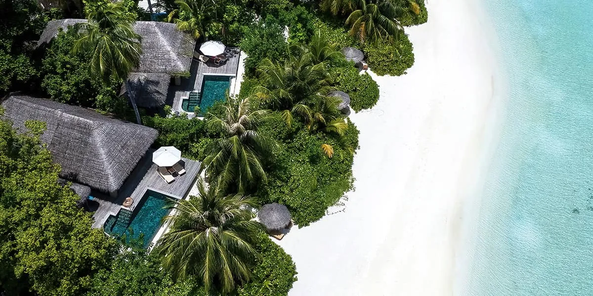 Aerial view of pool villas among palms beside a white sand beach