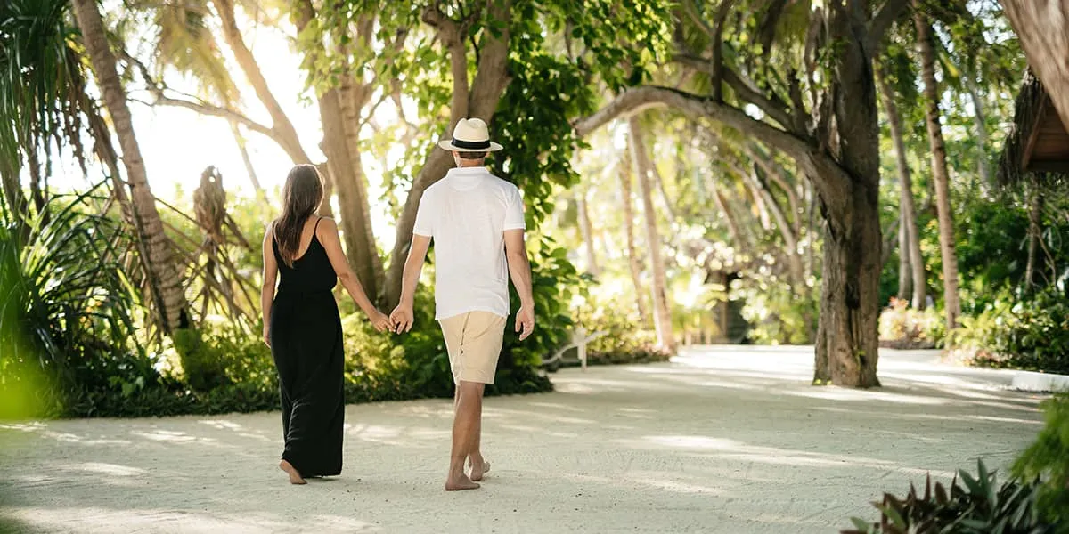 Couple walking hand in hand along a sandy path shaded by trees at Baros