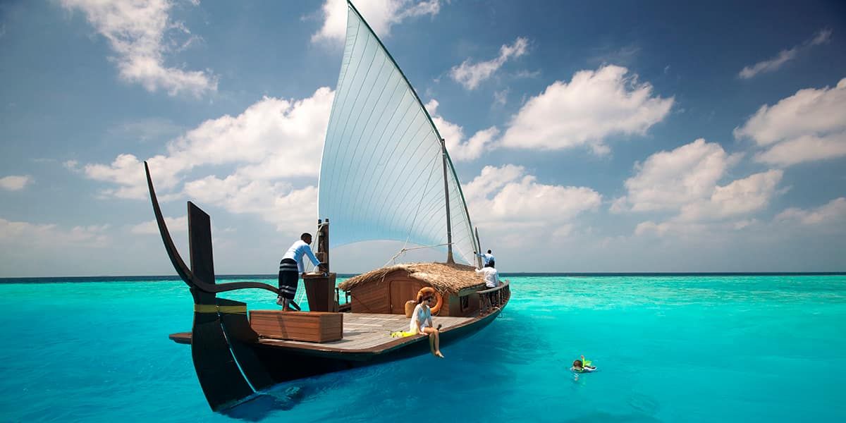 Traditional dhoni with a white sail anchored in a turquoise lagoon, snorkeller nearby