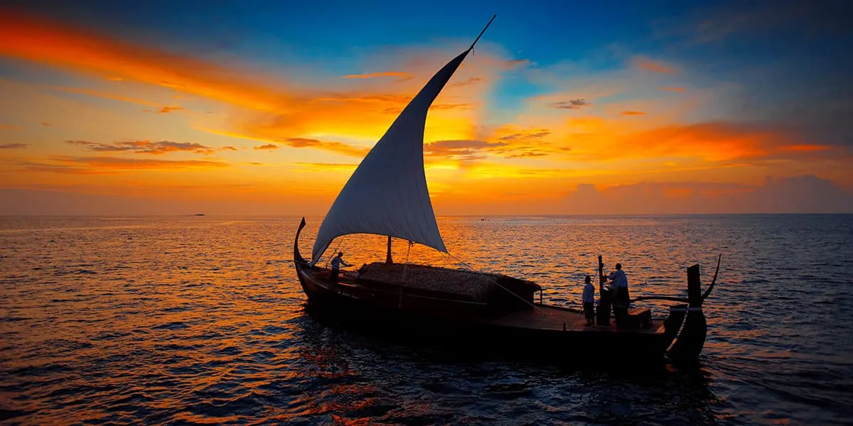 Traditional dhoni with a raised sail silhouetted against an orange sunset