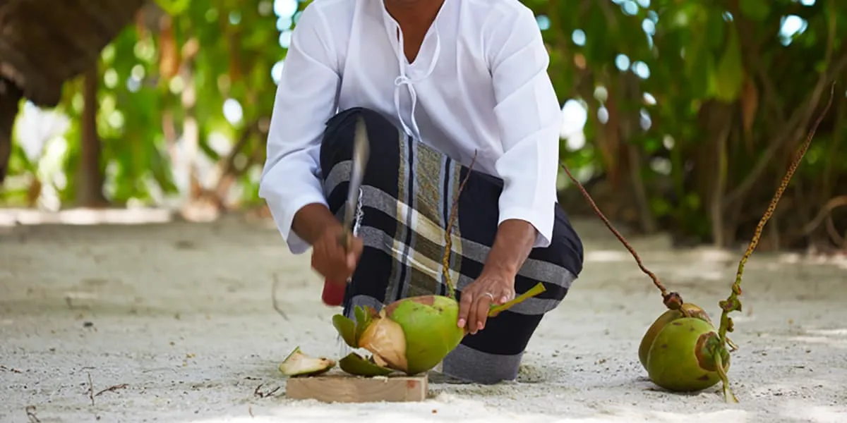 Man in a sarong cutting open fresh coconuts on the sand