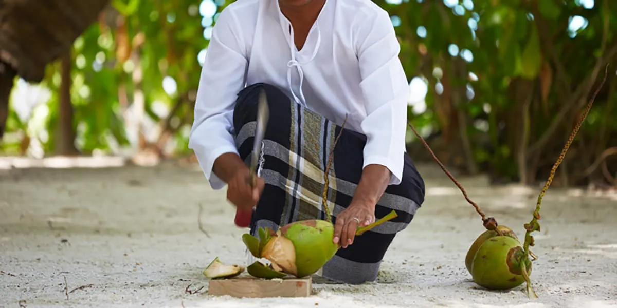 Man in a sarong cutting open fresh coconuts on the sand