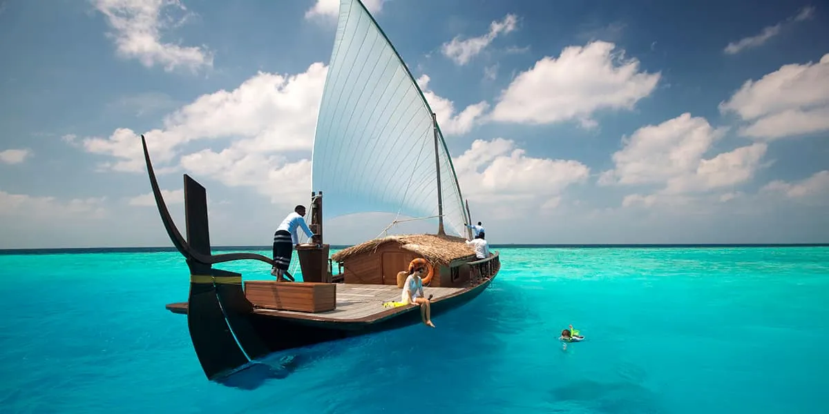 Traditional dhoni with a white sail anchored in a turquoise lagoon, snorkeller nearby