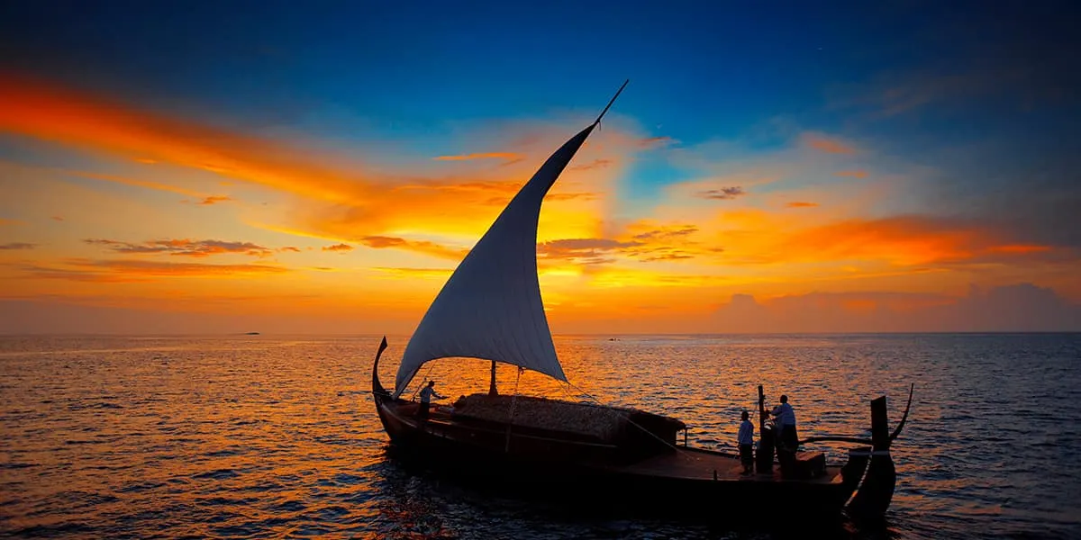 Traditional dhoni with a raised sail silhouetted against an orange sunset