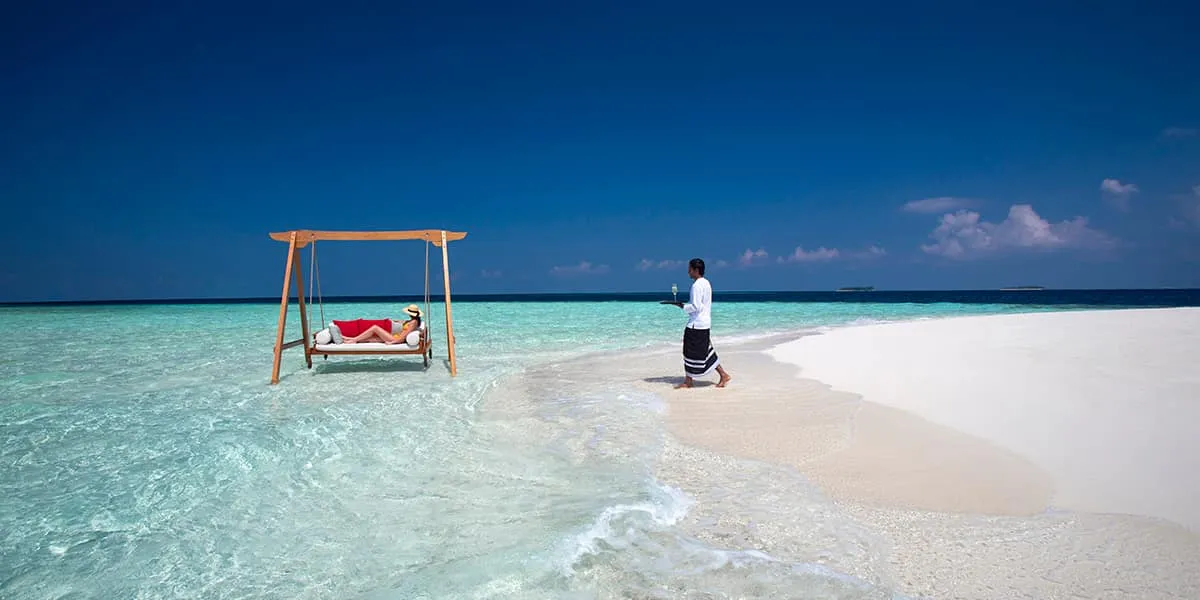 Guest on a swing daybed in shallow water off a sandbank, host walking nearby