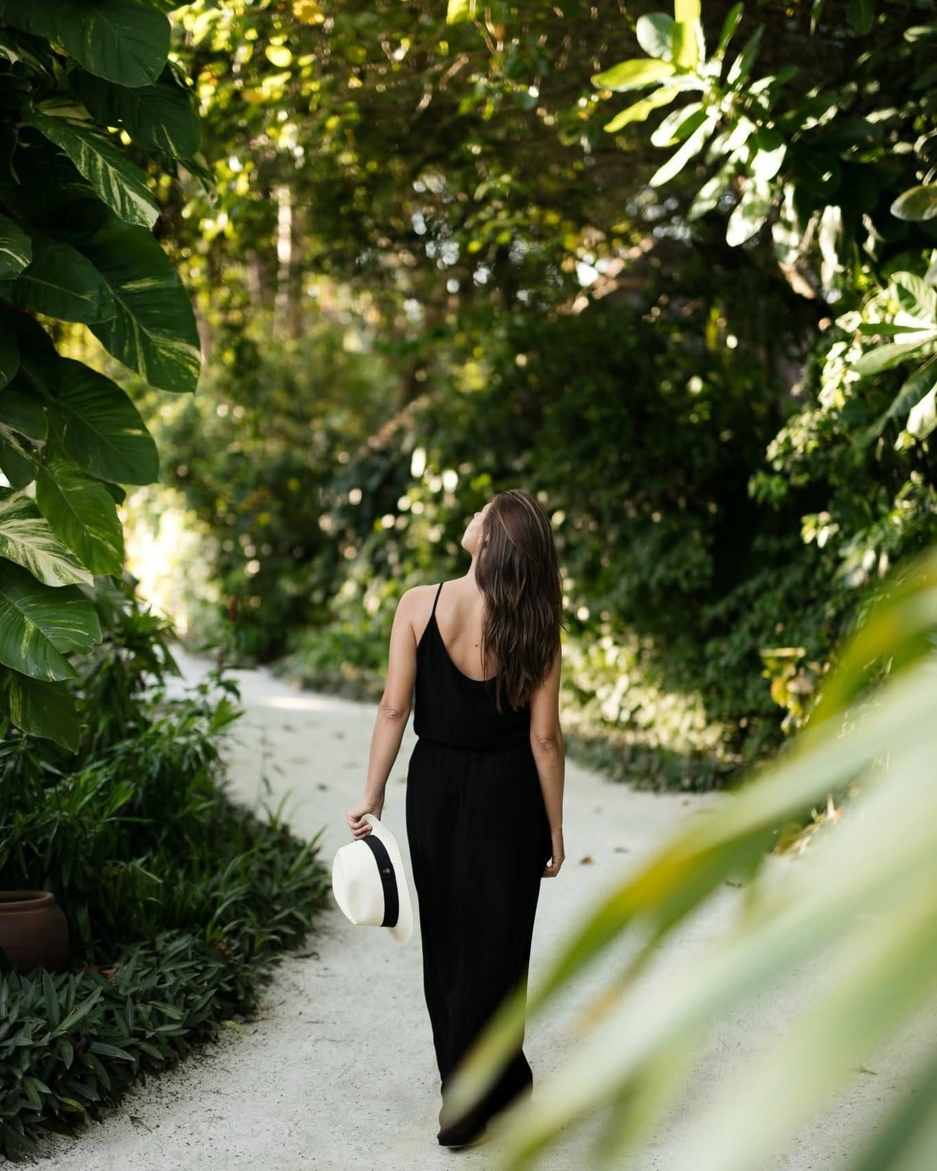 Woman in a black dress walking a sandy jungle path, seen from behind