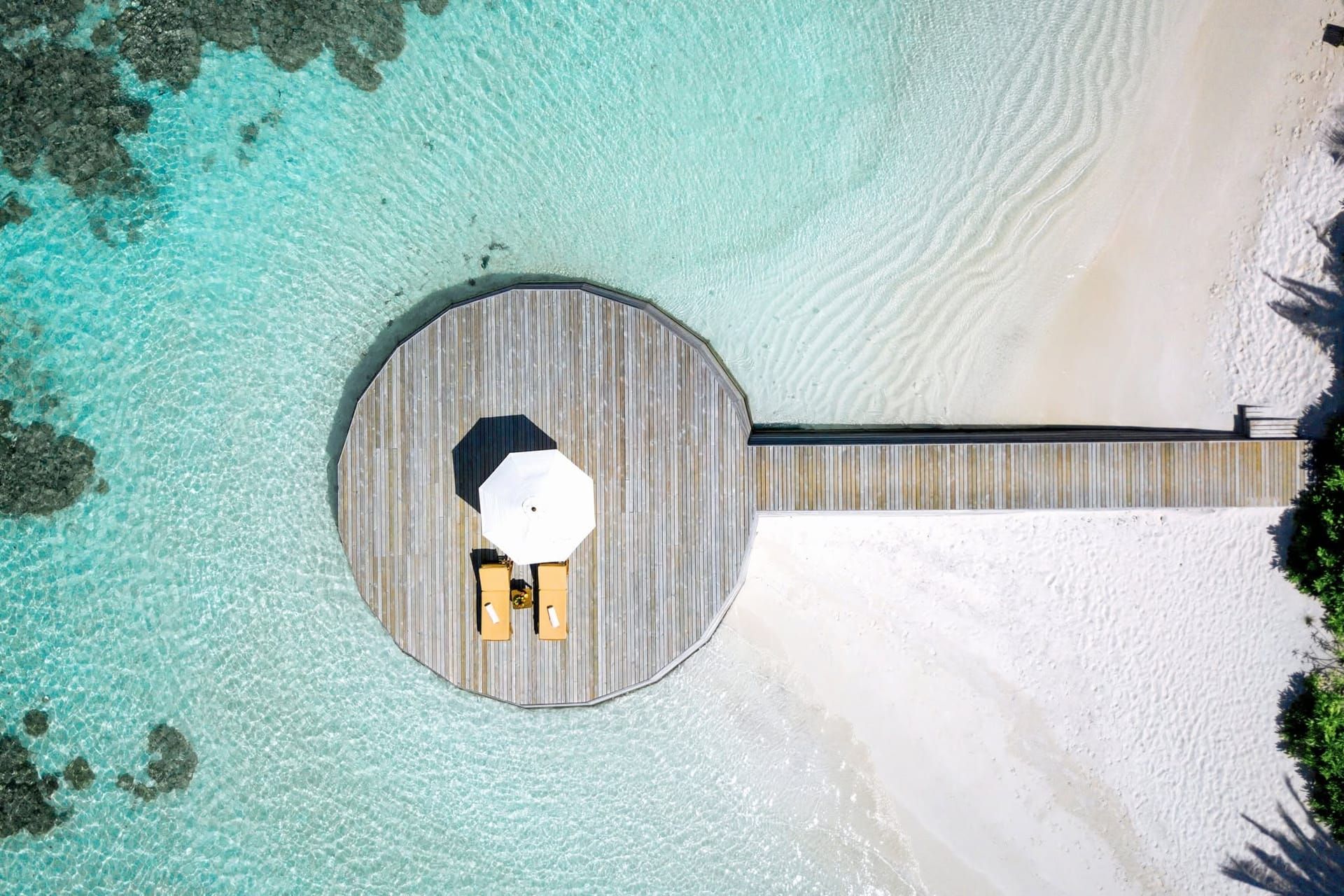 Aerial view of a circular jetty-end deck with a parasol over white sand and turquoise water