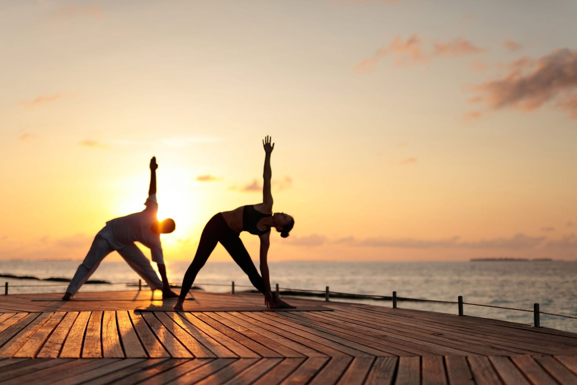 Two people holding a yoga triangle pose on a deck at sunset