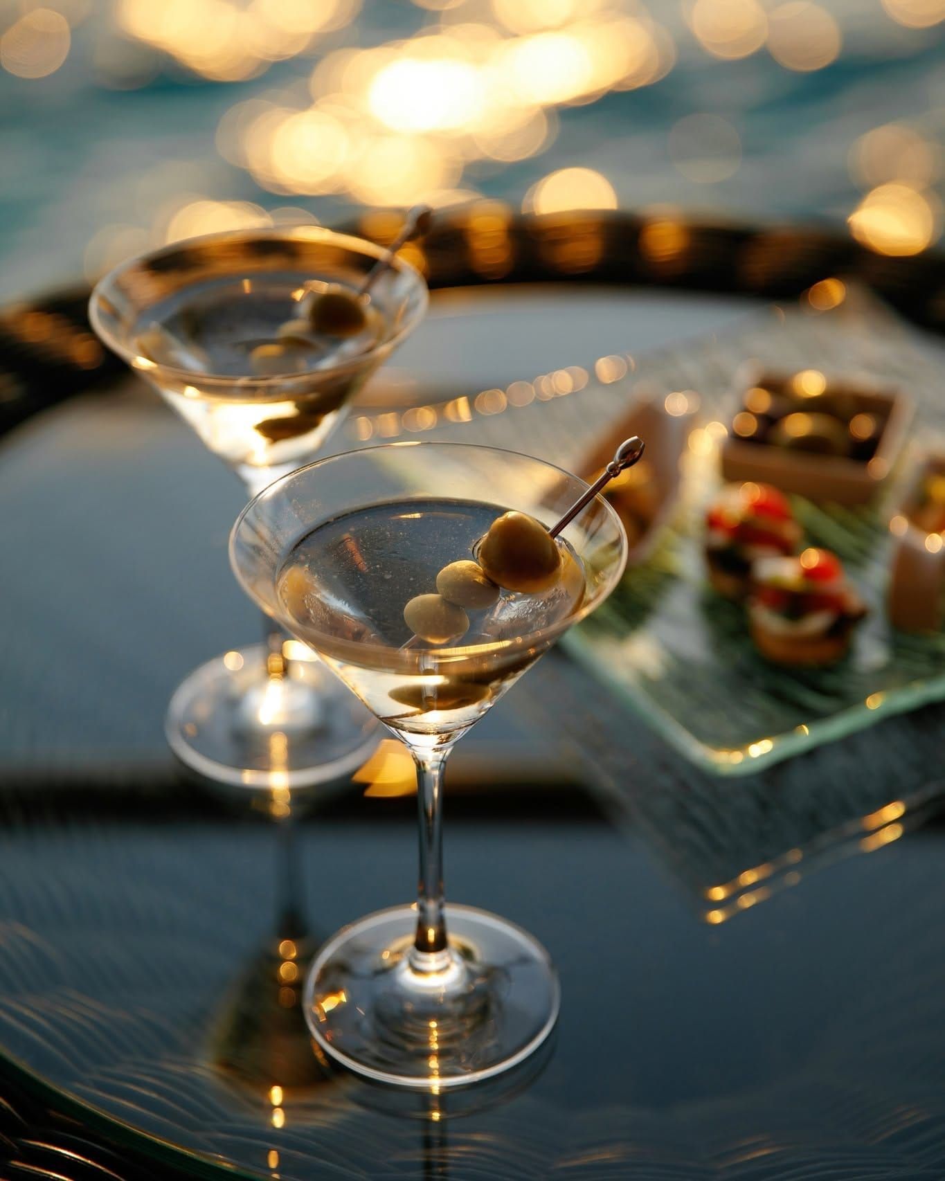 Martini glasses with olives and canapés on a tray against a glittering sunset backdrop