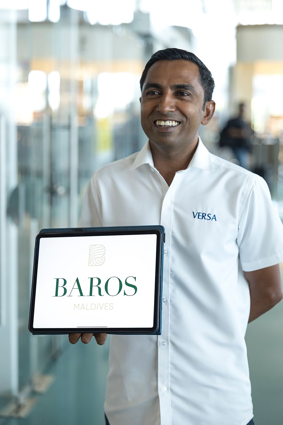 A smiling Versa Hospitality airport representative holding a tablet with the Baros Maldives welcome sign