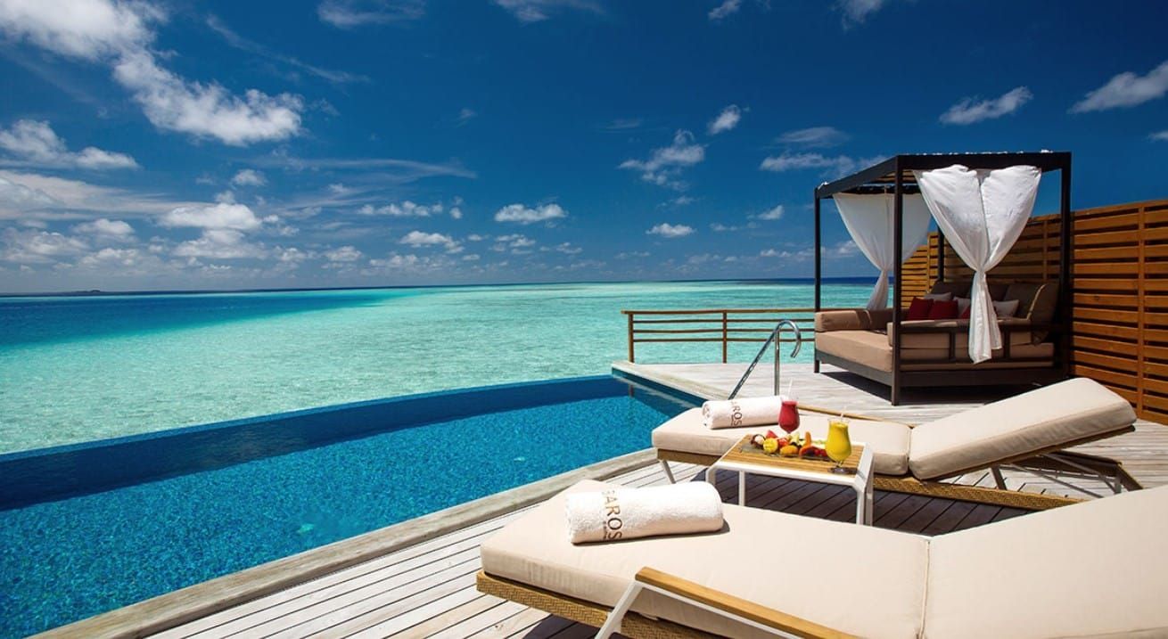 Overwater villa deck with infinity pool, cabana bed and loungers under blue sky