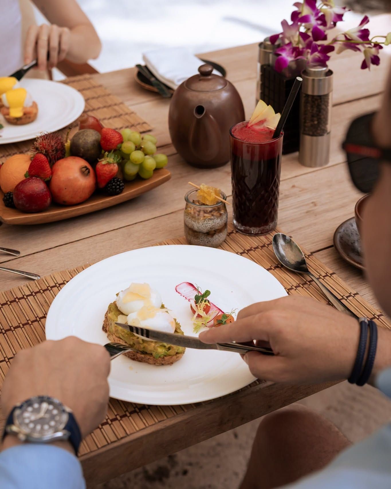 Hands slicing a breakfast dish at a table laid with fruit and orchids