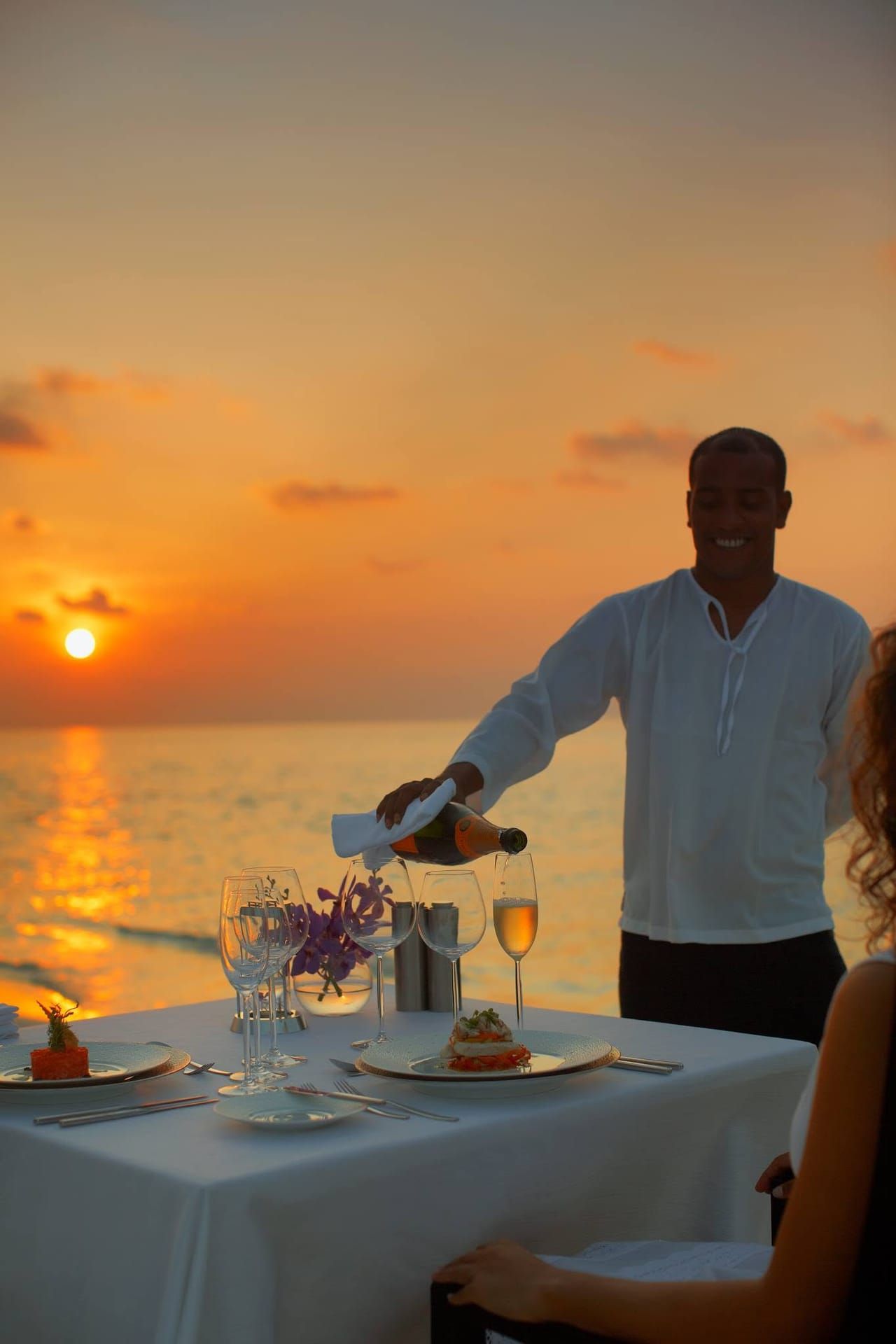Waiter pouring drinks at a table set for dinner on a deck at sunset