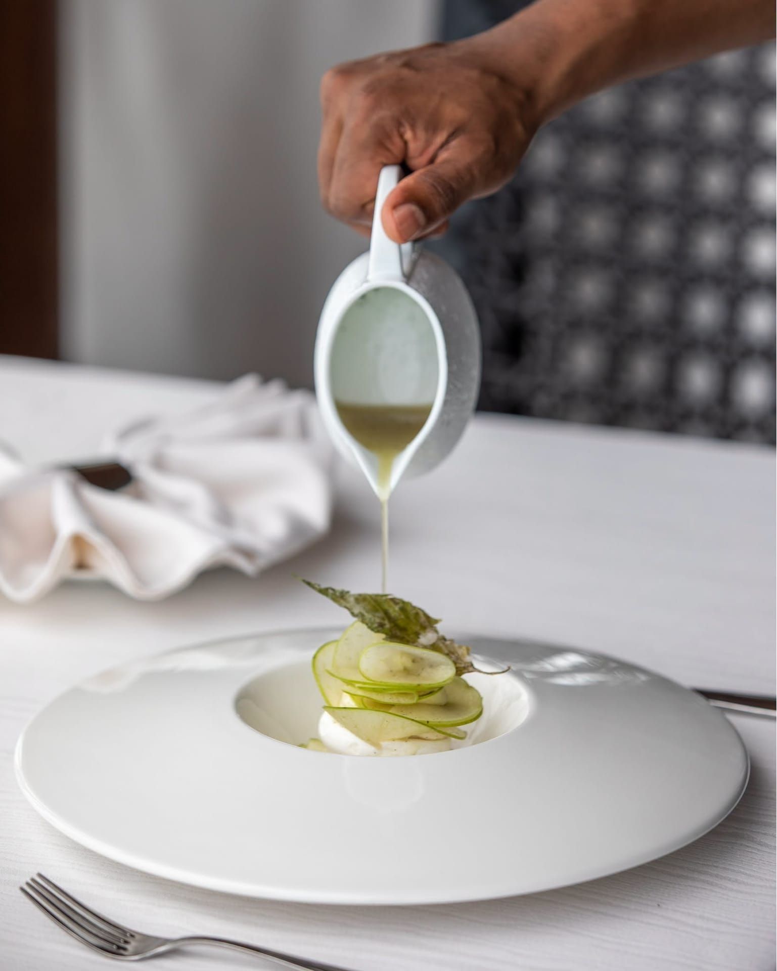 Server pouring sauce over a plated dish at a white-clothed table