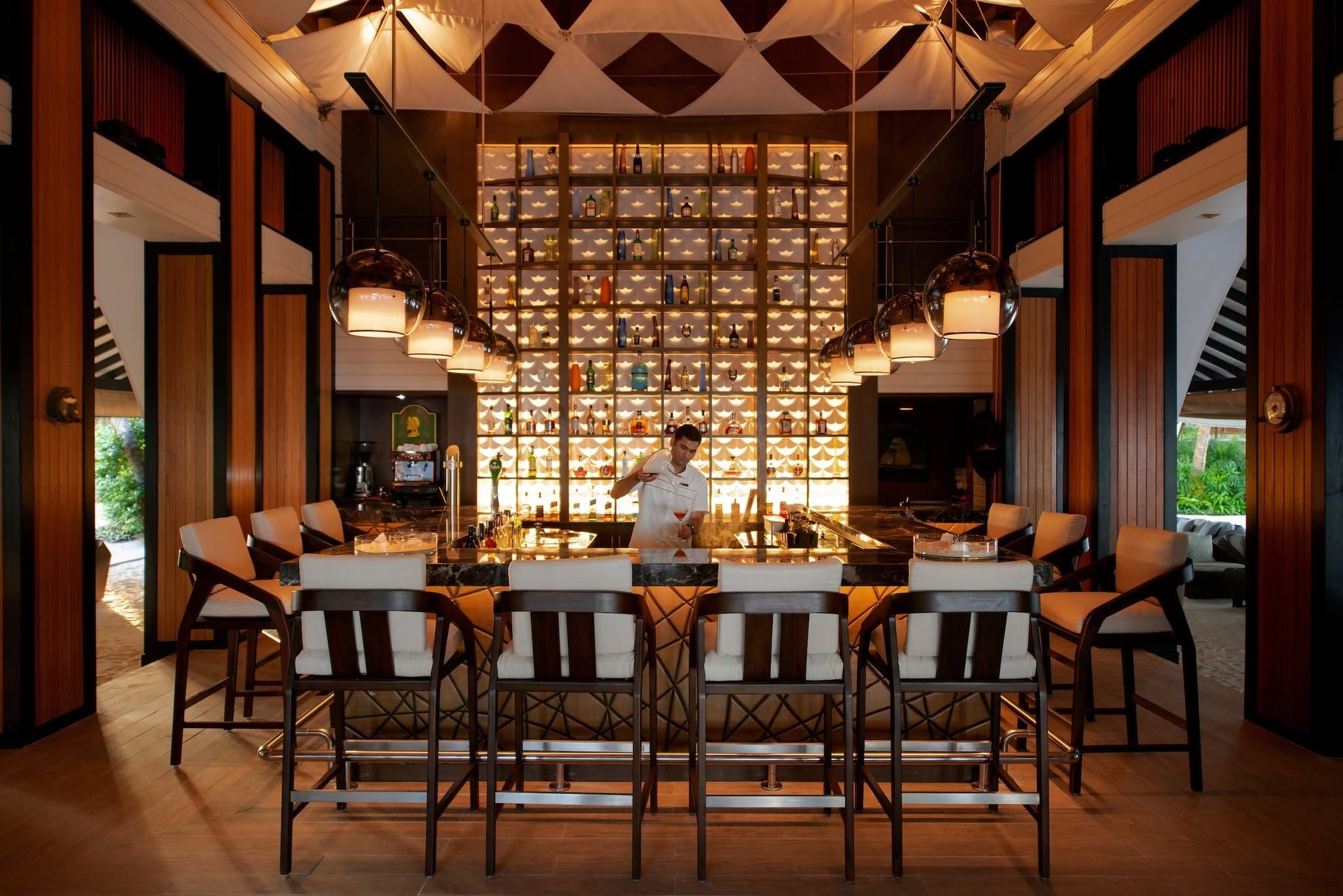 Bartender behind a backlit bar with rows of bottles and high bar stools