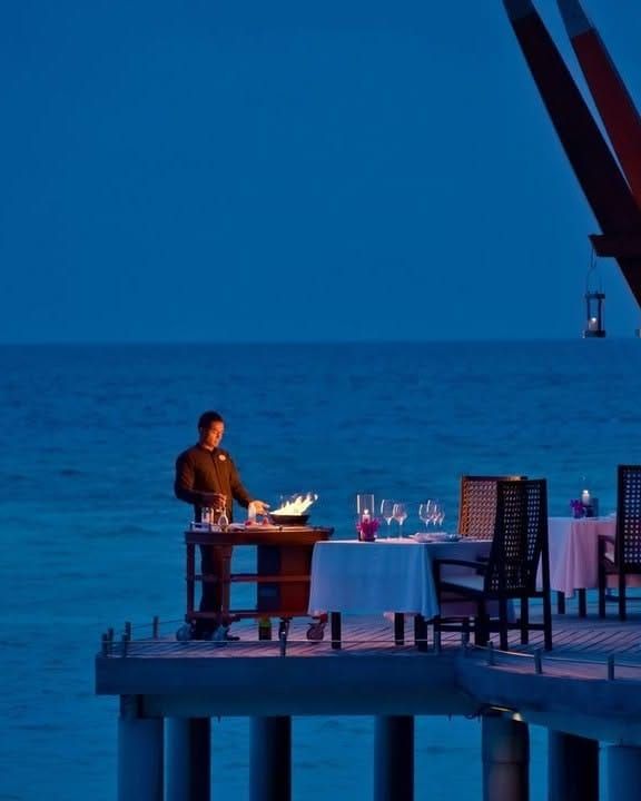 Waiter at a table set for dinner on a jetty over deep blue water at dusk