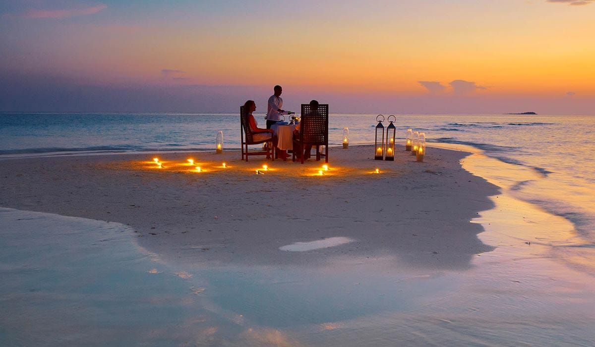 Couple at a candlelit table on a sandbank at sunset, attended by a waiter