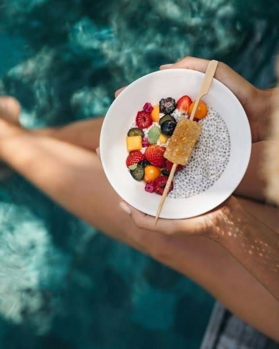 Hands holding a bowl of chia pudding topped with fresh fruit above clear water