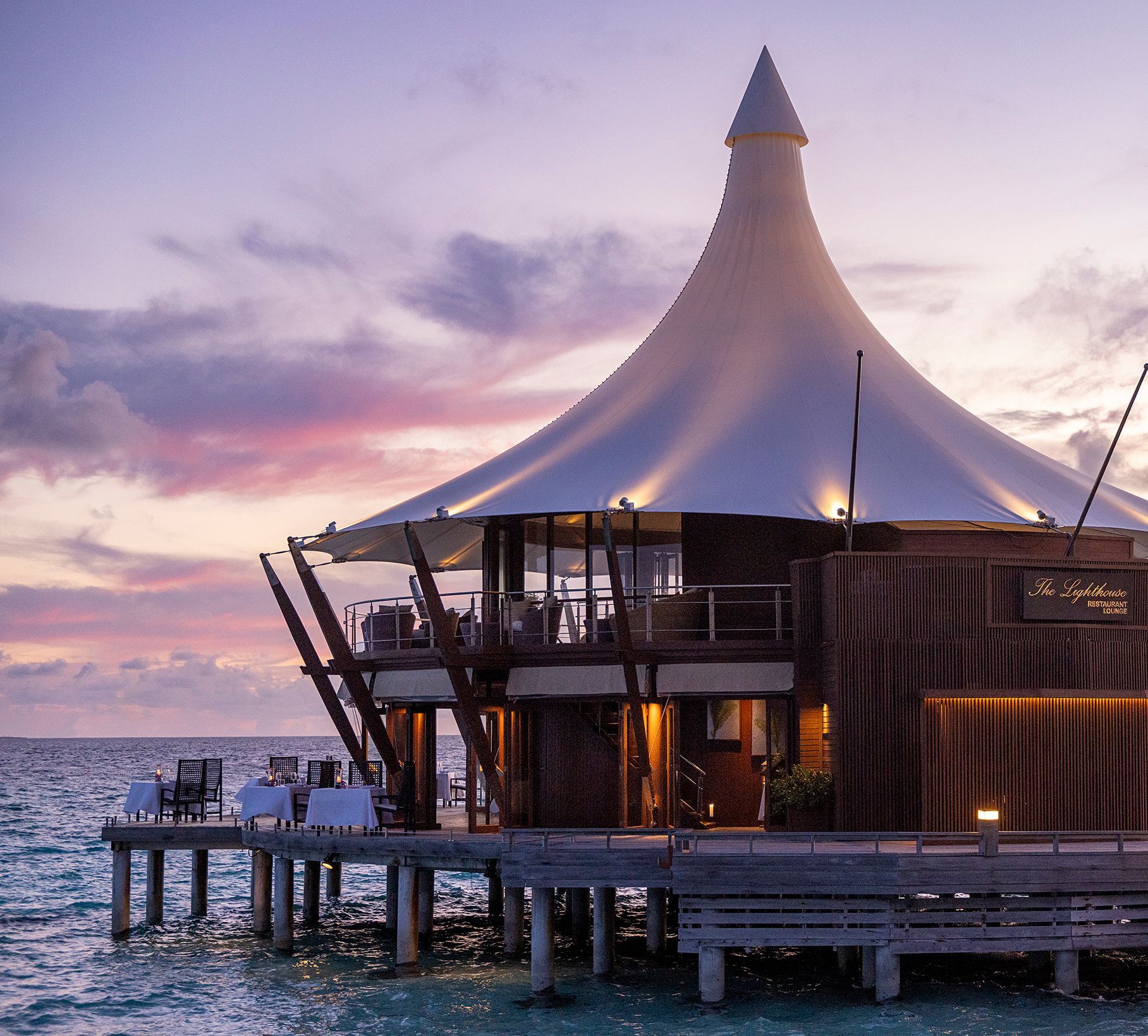 The Lighthouse restaurant on stilts over the lagoon at sunset, its white canopy glowing against a pink and gold sky
