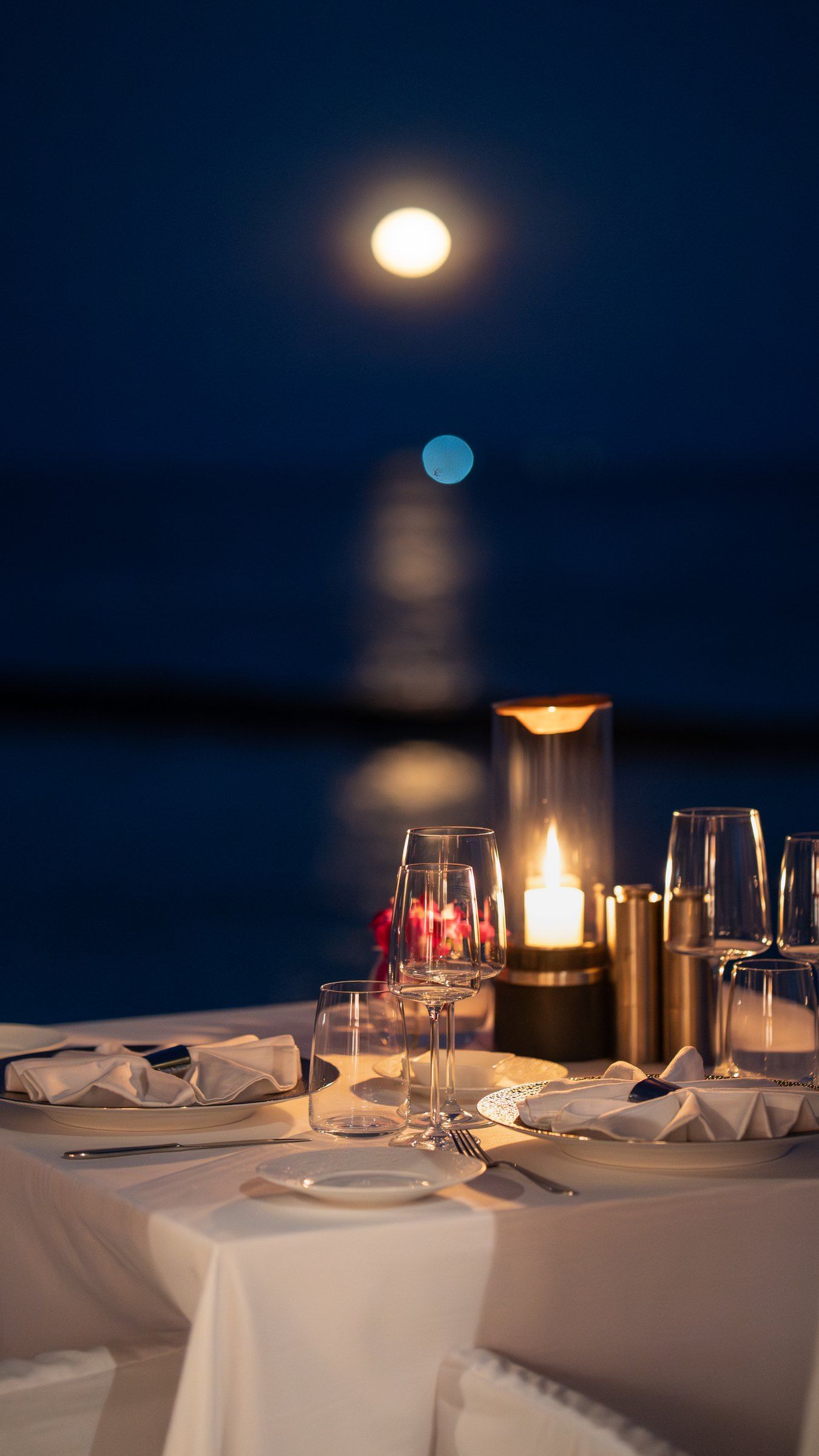 A candlelit table set for dinner by the water under a moonlit sky
