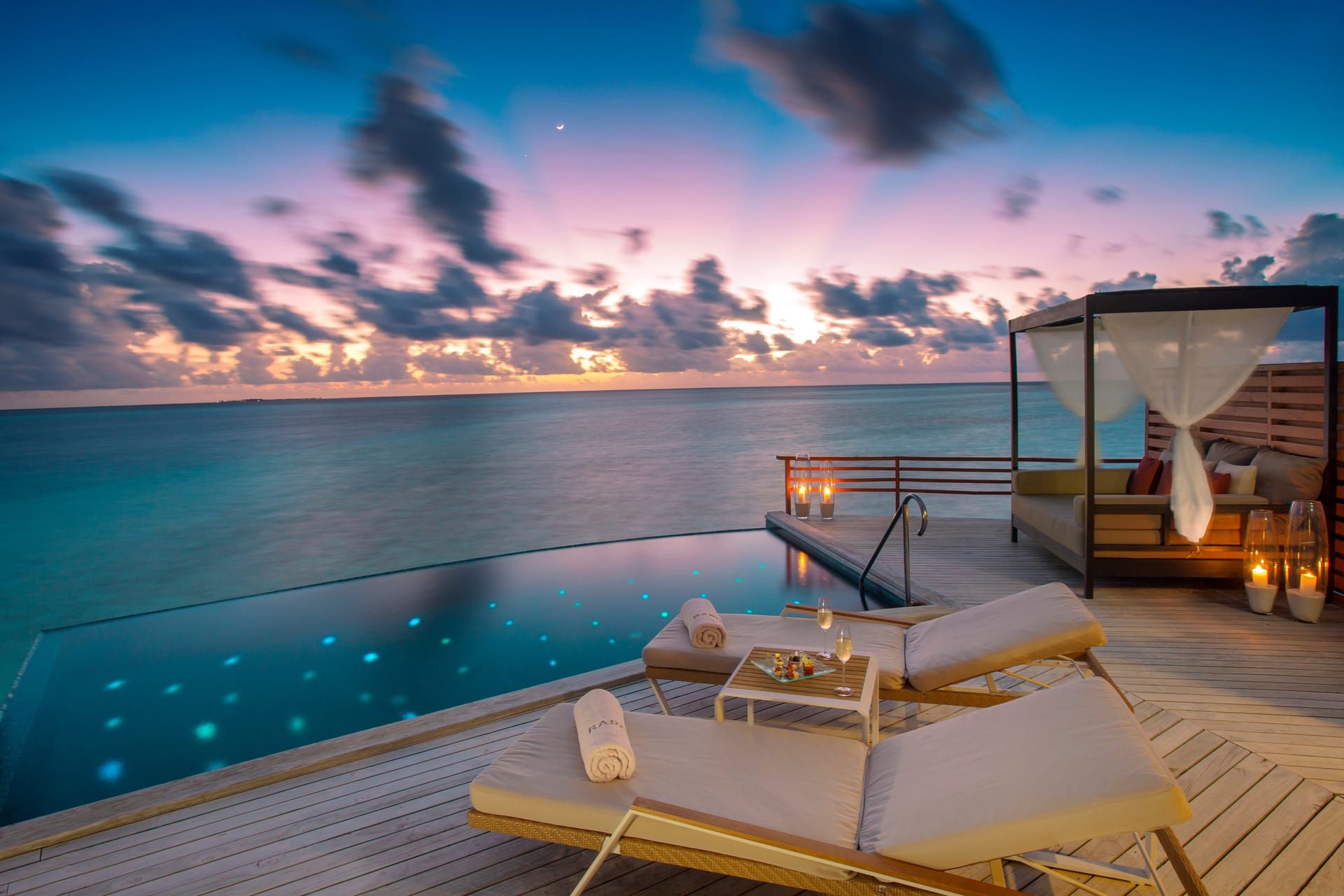 Overwater villa pool deck with cabana and loungers at dusk, pool lights glowing
